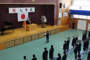 体育館で舞台上の校長先生に向かって、舞台下の生徒が新入生代表挨拶を行っています。