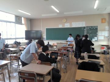 2年5組の教室内で、複数の生徒と教員が、机に座ったり、立ったりしています。黒板と大きな窓と大型テレビが見えています。