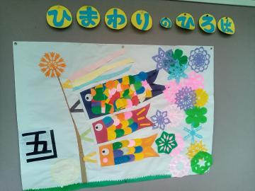 色とりどりの鯉のぼりと花の飾りが並んだ学校の掲示板で上部にひまわりの文字があり左側に大きな五の文字がある