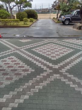 幾何学模様の石畳の通路が手前に広がり右側に黒い車が駐車され奥に低い植栽と道路が見える