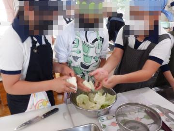 複数の人がエプロンと帽子を着用して大きなボウルで葉物野菜を手でちぎっている調理場面