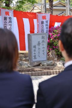 後ろ姿の二人が石碑と赤白の垂れ幕が並ぶ式典会場を見つめている