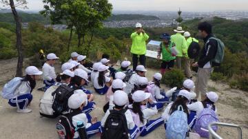 児童たちが山の斜面で座って引率の大人から説明を受けているようす