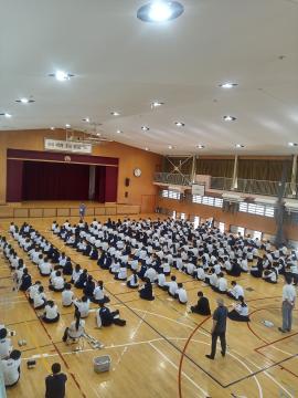 体育館で床に整然と座る多数の生徒と壇上の舞台を見下ろす高所からの全景