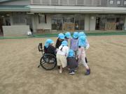 校庭で複数の子どもが帽子をかぶって車いすを囲んでいる