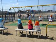 公園の砂地のそばのベンチに帽子をかぶった子どもたちが数人座り背景にブランコと運動場の風景