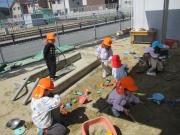 園庭でオレンジの帽子をかぶった子どもたちが砂場や道具を使って遊んでいる