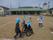 校庭で青い帽子をかぶった子どもたちが車椅子の周りで遊んでいる様子