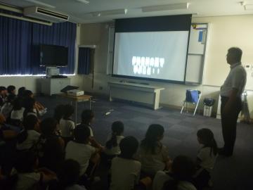 暗くした教室で床に座る児童たちが前方のスクリーンに映る映像を見ている傍らで教師が立っている
