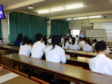 教室で教員が立って説明をし、生徒たちが机に座って聞いている風景