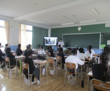 教室で黒板とモニターを前に座席に着いた生徒たちが授業を受けている様子