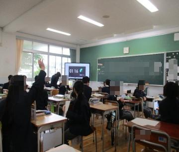 窓のある教室で黒板と時計の前に机が並び制服を着た生徒たちが着席して数人が手を挙げている