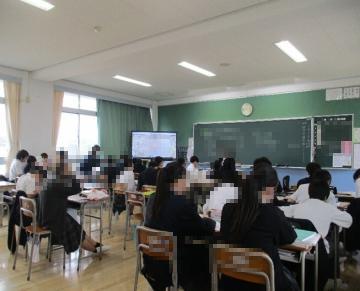 教室で多くの生徒が机に座って前方の黒板とディスプレイを見ている様子