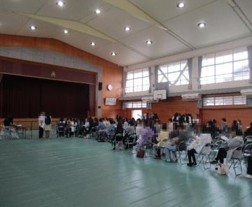 体育館の室内で天井の高い会場に椅子が並べられ多くの人々が座ったり列を作って受付や待機をしているイベントの様子