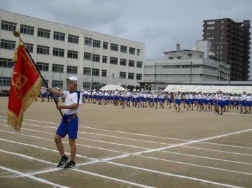 運動場で旗を持つ生徒が先頭に立ち多数の生徒が列を作って整列している