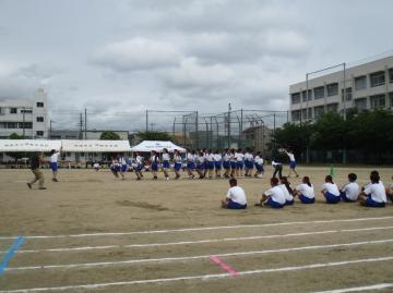 学校の運動場で屋外のグラウンドに生徒たちが整列し応援やクラスで長縄を跳んでいる様子