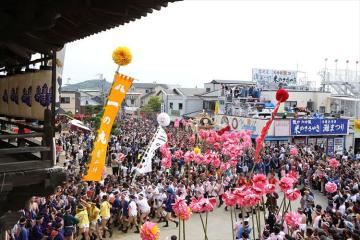 屋外の広場に大勢の人々が集まりカラフルな風船や長い吹き流しが飾られた祭りのパレードが行われている