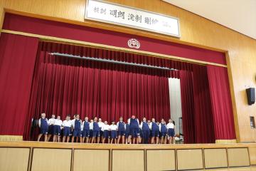 学校の講堂の舞台で制服を着た生徒たちが横一列に並んで立っている