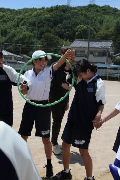 屋外の運動場で体操服の学生たちが輪を持って一人の学生の頭上にくぐらせる遊びをしている