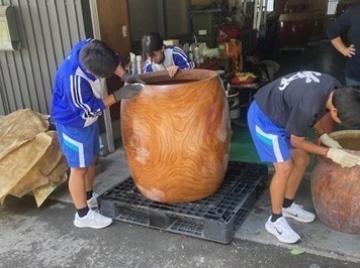 作業服の学生たちが大きな木製の樽を屋内の作業場で磨いたり手入れしたりしている様子