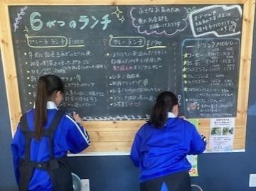 大きな黒板に書かれたランチメニューを前に青いジャージを着た二人が黒板に向かって書き込んでいる