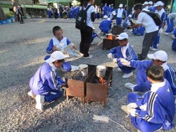 屋外で青い運動着と白い帽子を着た学生たちが金属のコンロで鍋を温めたり食事を準備している様子