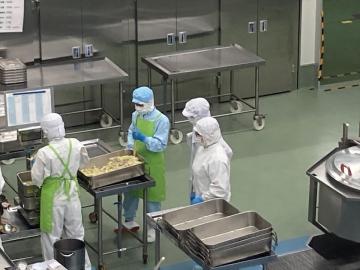 食品加工の作業場で白い防護服とマスクと青い手袋を着けた作業員がトレイの製品を整理検査している場面