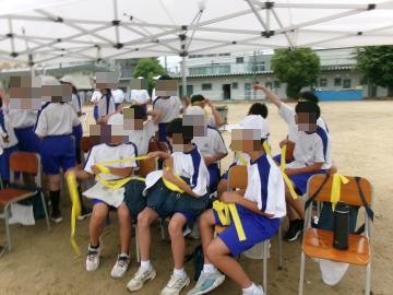 運動着を着た生徒たちが校庭のテントの下で椅子に座りタオルやゼッケンのような黄色い布を手にしています。
