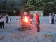 夕暮れの屋外の広場で数人が木製の台に積まれた焚き火を囲んでいる光景