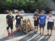 屋外の砂利敷きの場所で数人の人々が木材で組んだまきの前に並んでいる