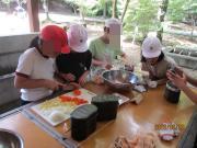 屋外のテーブルで帽子をかぶった子どもたちがボウルやまな板を使って野菜を切り食事の準備をしている