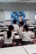 教室でスクリーンに資料を表示している講師が前に立ち生徒が机に向かって座って聴いている場面の写真