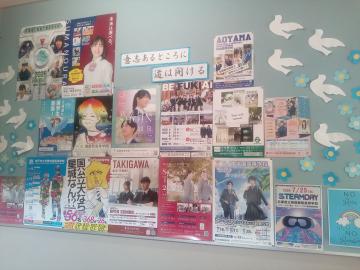 壁に貼られた複数のイベントや告知のポスターとチラシが並び背景に白いハトの切り絵が飾られている掲示板の写真