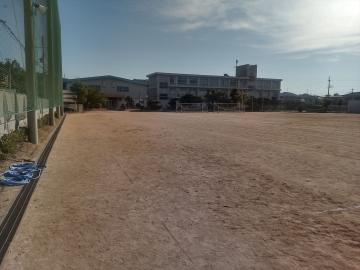 広い土の校庭の奥に校舎と旗が立ち青空の下に広がる学校の風景