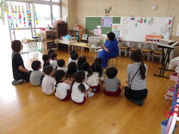 室内で4歳児がお話の先生の絵本の読み聞かせを聞いている様子