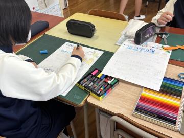 教室の机に座り学習資料に書き込む生徒とカラーペンや筆箱が並ぶ様子