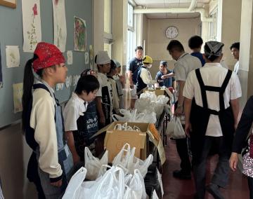 学校の廊下で複数の生徒と参加者が白いレジ袋や段ボール箱に入った物資を並べて配布している様子