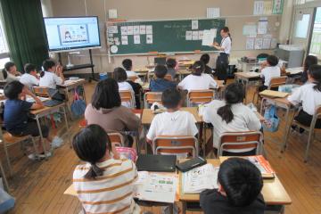 教室内で、5年生の児童が外国語の授業で先生の説明を聴いている様子
