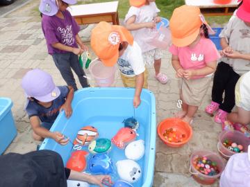 3歳児や5歳児の子供たちが、屋外で、水を張った大きなプラスチック容器の中でおもちゃの魚やボールを浮かべて、すくって遊んでいる様子