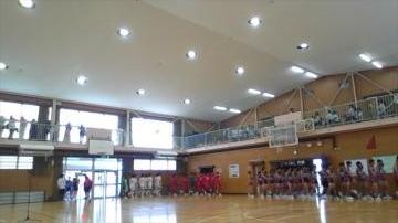 屋内の体育館でユニフォームを着た人々が床の両側に整列し二階の観覧席に観客がいる様子