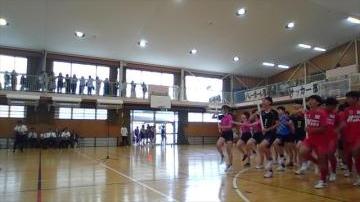 体育館の床に整列した複数の学生が赤や青のユニフォームを着て並び観客が上の観覧席から見守っている