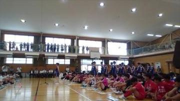 体育館の中で色とりどりのユニフォームを着た多くの子どもたちが床に座って並び上の観覧席に人々が立っている行事の様子