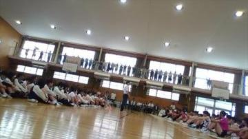 体育館で床に座っている多数の児童生徒と上階の手すりに沿って立つ人々が集まっている様子