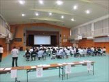 体育館で高等学校の先生のお話を聞く生徒と保護者の様子