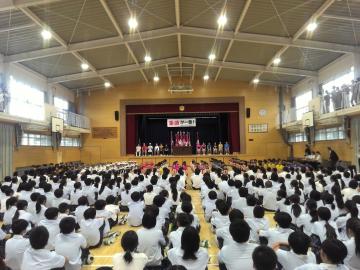 体育館の舞台で発表が行われているのを床に整列して座って見ている全校生徒