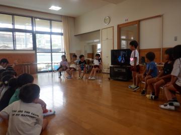 テレビの画面に写真を写し、座っている子供たちに写真の説明をしている5歳児の子供