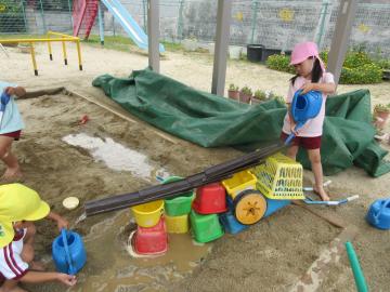 砂場でバケツを重ねて樋をつなげ、ジョウロで樋に水を流す子供