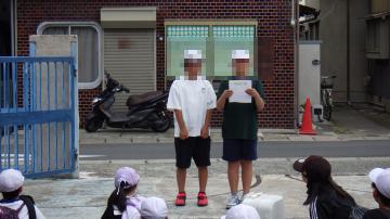 正門の前で児童2名が修学旅行出発のあいさつをしている様子