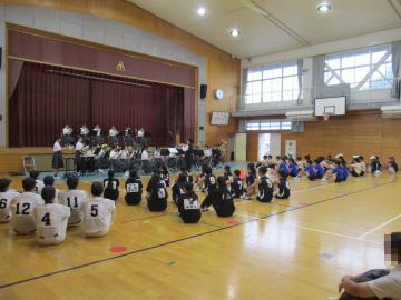 体育館の床にユニフォームを着た複数の生徒が整然と座り舞台上で数人の演奏者が演奏している光景