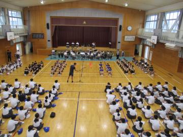 体育館のステージ前に座る部活動の生徒と床に整列して座る多数の生徒が集まる集会の様子